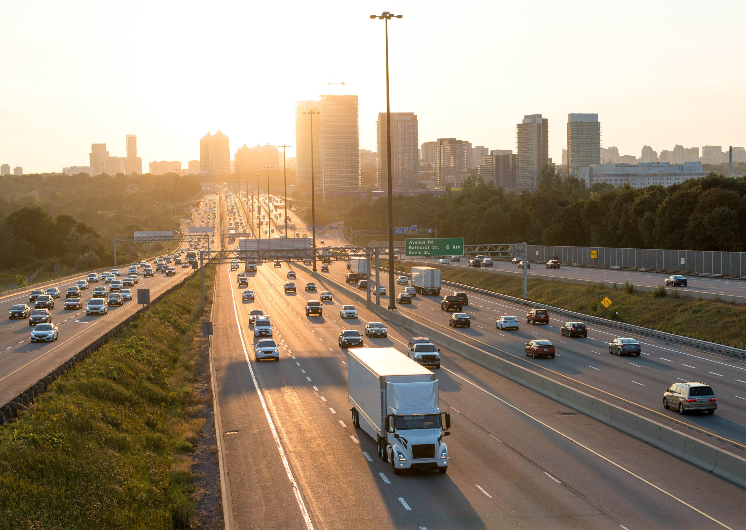 Busy multi-lane highway with cars and trucks moving in both directions. Sun rises or sets behind a distant city skyline, with trees lining the roadside.