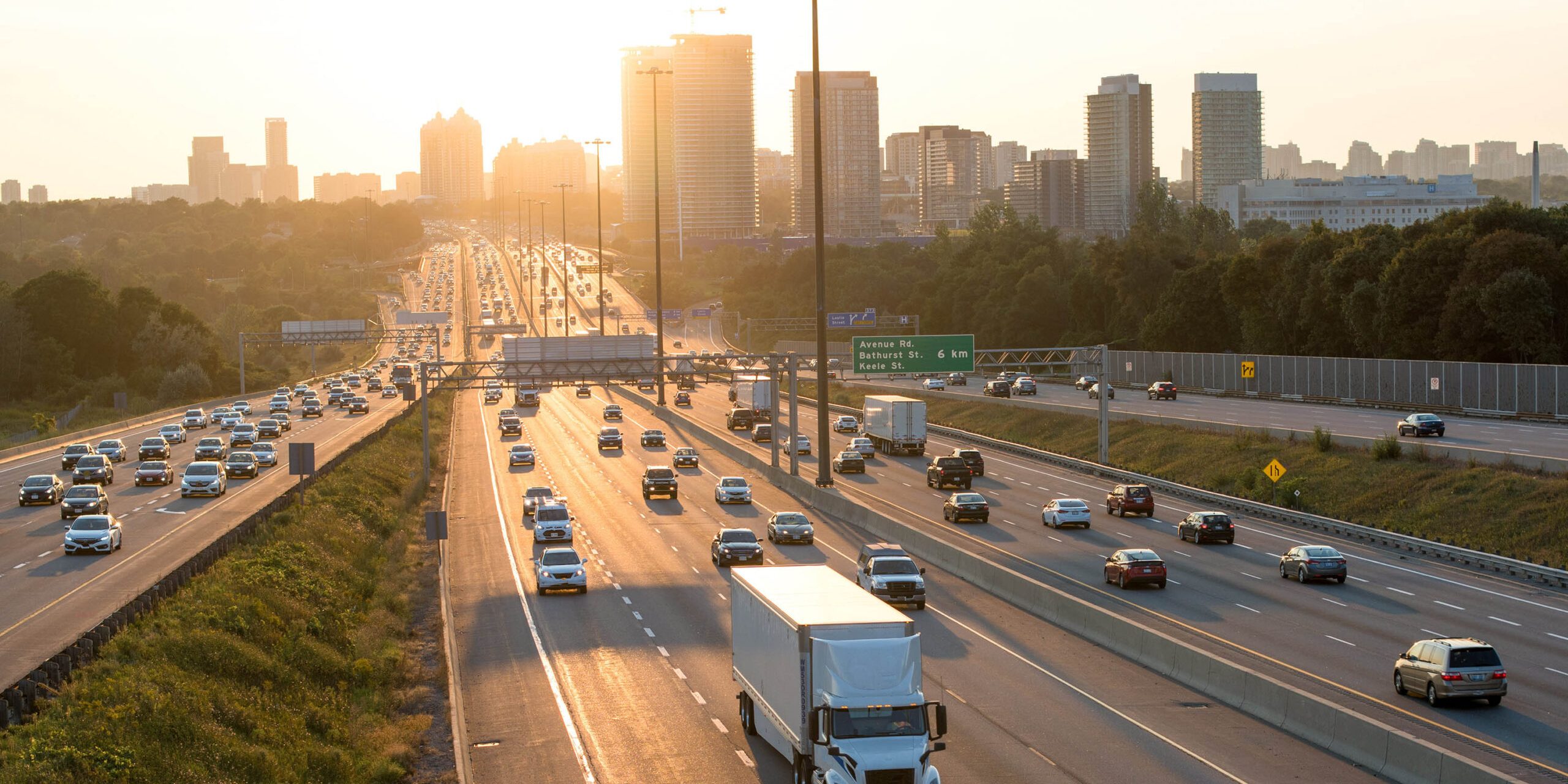 Busy multi-lane highway with cars and trucks moving in both directions. Sun rises or sets behind a distant city skyline, with trees lining the roadside.