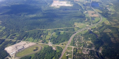 Aerial view of a landscape with a highway running through forested areas, open fields, residential zones, and an industrial area featuring a circular structure.