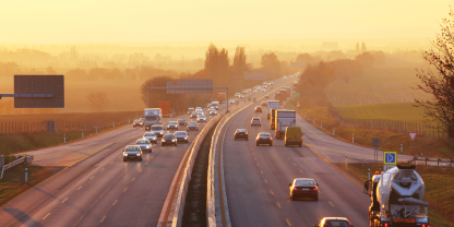 A highway with multiple lanes in each direction, carrying cars and trucks under a golden sunset or sunrise. The road is surrounded by open fields and distant trees.