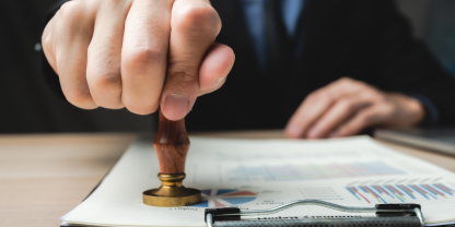A person in formal attire stamps a document clipped to a clipboard. The document contains colorful charts or graphs, suggesting official approval or review.