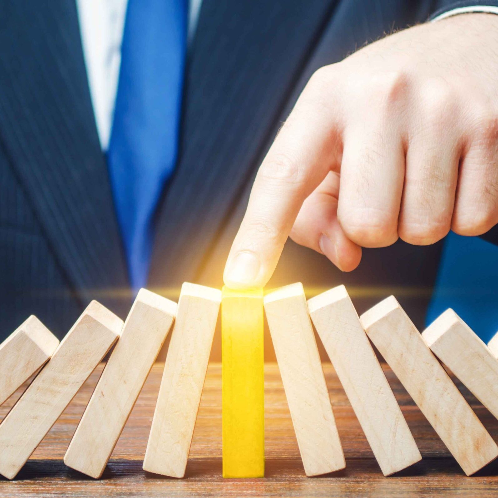 Une personne en costume et cravate bleue utilise son index pour arrêter une rangée de dominos en bois en train de tomber. Le domino touché est mis en évidence en jaune pour souligner l’action.